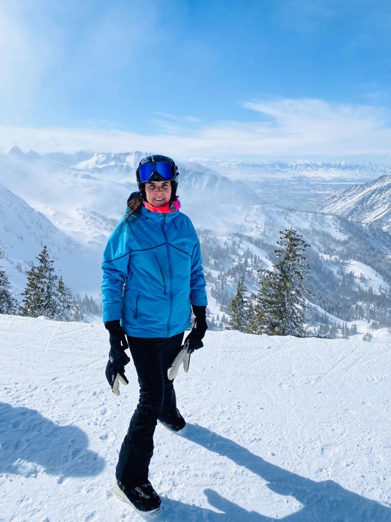 Liz Healy in a ski helmet, goggles, and winter jacket, standing on a snowy mountain with a mountain range in the background.