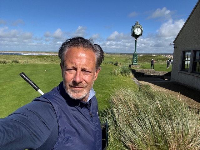 A man in a navy vest takes a selfie on a golf course.