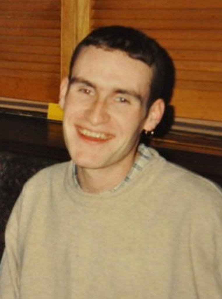 A young man with short dark hair, wearing a beige crew-neck shirt over a plaid button-up, smiles at the camera.