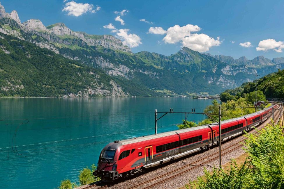 一列鮮紅色的ÖBB列車沿著波光粼粼的碧綠湖畔行駛，背景是雄偉的奧地利群山與藍天白雲，展現歐鐵奧地利通行證的風景鐵道冒險。