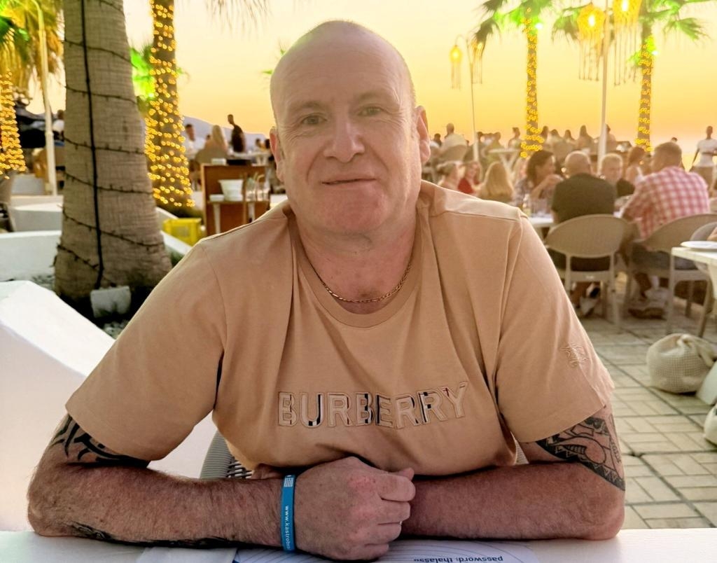 A man with short hair and tattoos on his arms, wearing a light brown shirt, sits at a table outdoors in front of palm trees and other people.