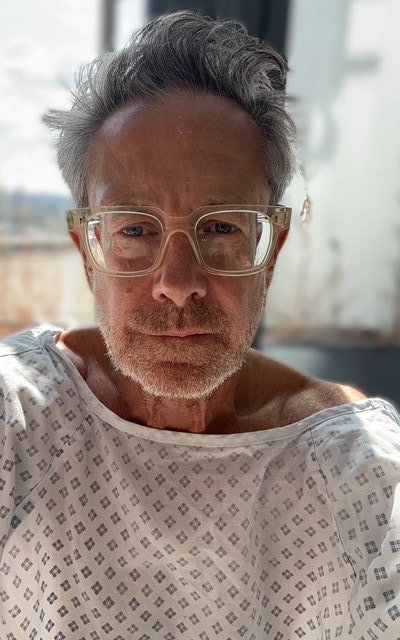 A man in a hospital gown and glasses looks directly at the camera.