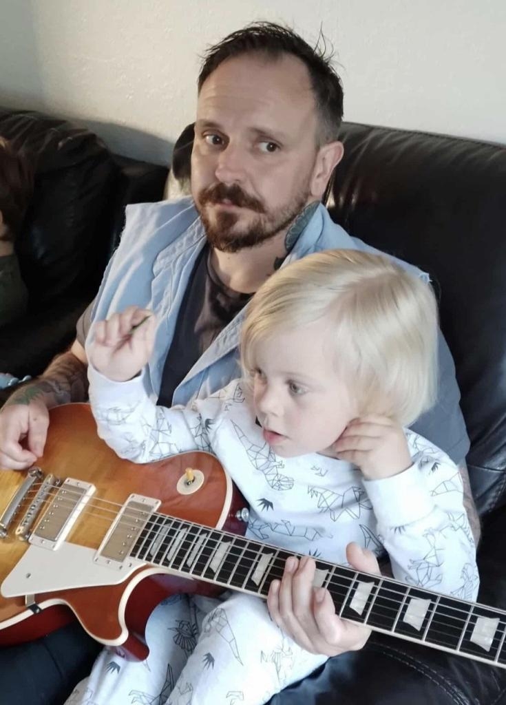 Mal Nash holding a young child with blonde hair who is holding an electric guitar.