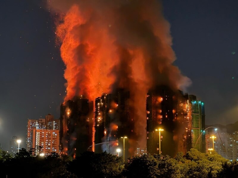 A large fire engulfs multiple high-rise buildings at Hong Fook Court in Tai Po, with thick orange flames and smoke billowing into the night sky, highlighting the severity of the blaze and potential concerns about insurance claims and rebuilding costs.