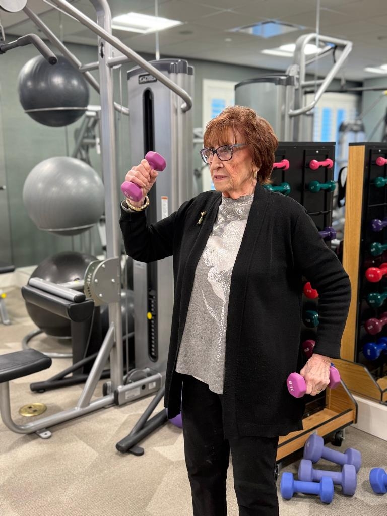 99 歲的雪莉·戈德曼雙手各持一個 3 磅重的紫色啞鈴，站在健身房裡，背景是健身器材。