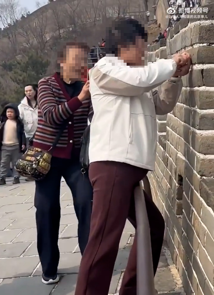 一名身穿白色上衣的中年女子在八達嶺長城的城牆上刻字，身旁一名身穿彩色條紋上衣的中年女子則用手機拍攝，疑在記錄同伴刻字的過程。（網上影片截圖）